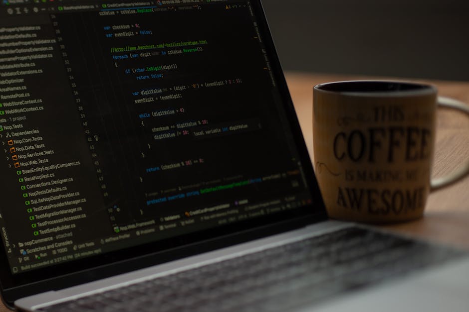 A close-up view of a laptop displaying lines of code on its screen, with a black background and colorful syntax highlighting. To the right of the laptop, there is a coffee mug with a handle, featuring