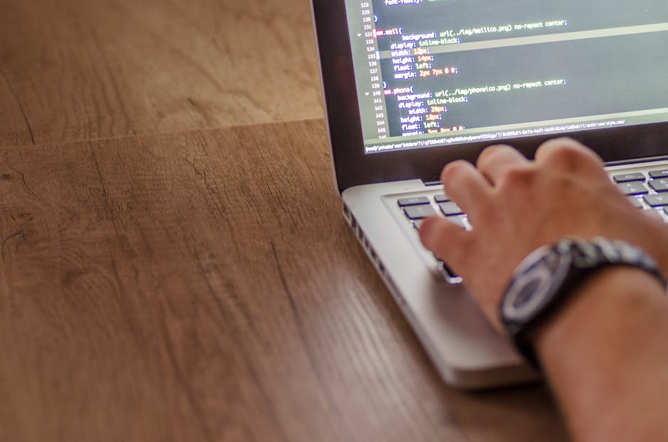 A close-up view of a laptop displaying lines of computer code with a dark background and colorful syntax highlighting, placed on a wooden desk. Next to the laptop, there is a ceramic coffee mug with t